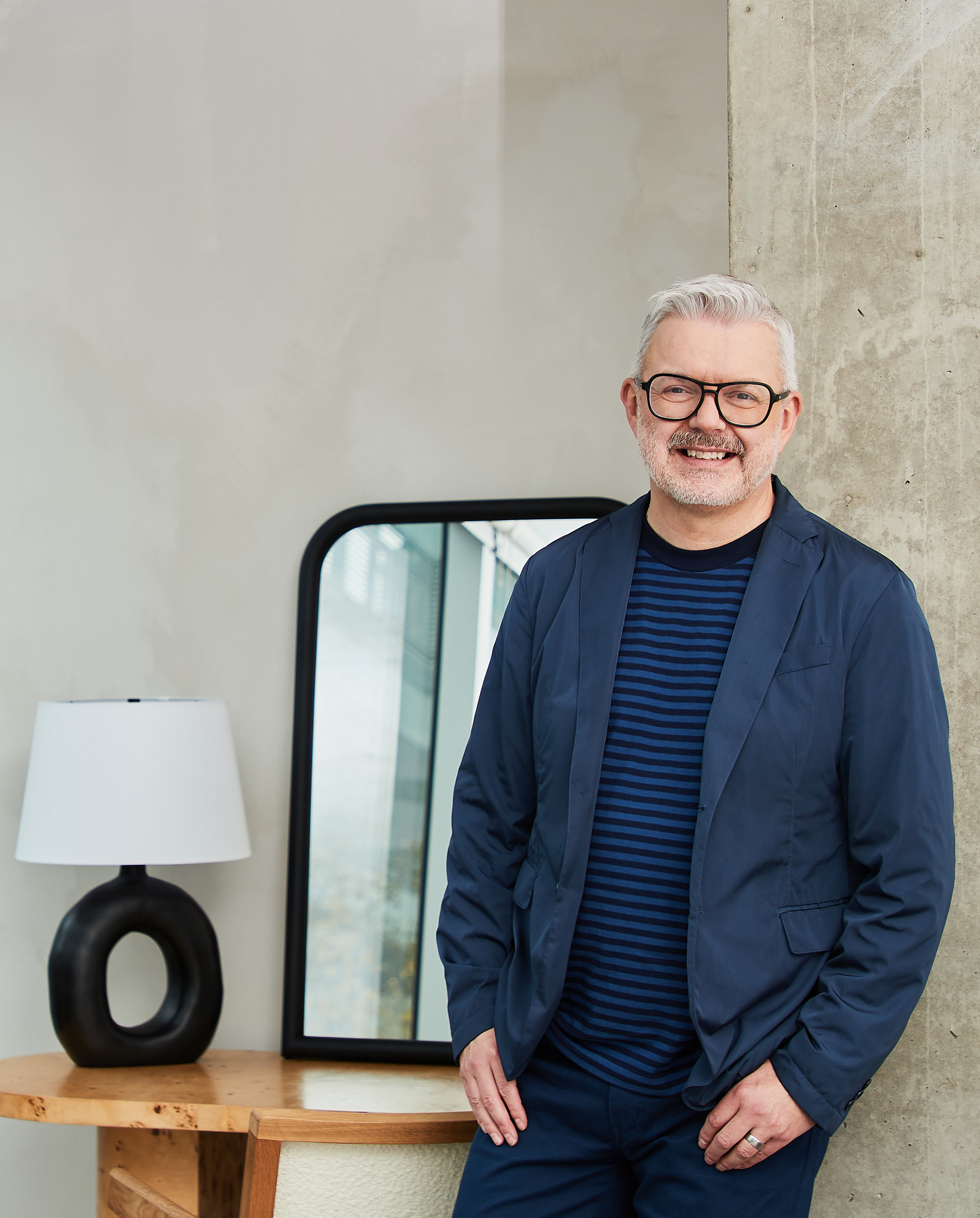 Man in a navy blazer standing beside a mirror and lamp, lifestyle portrait