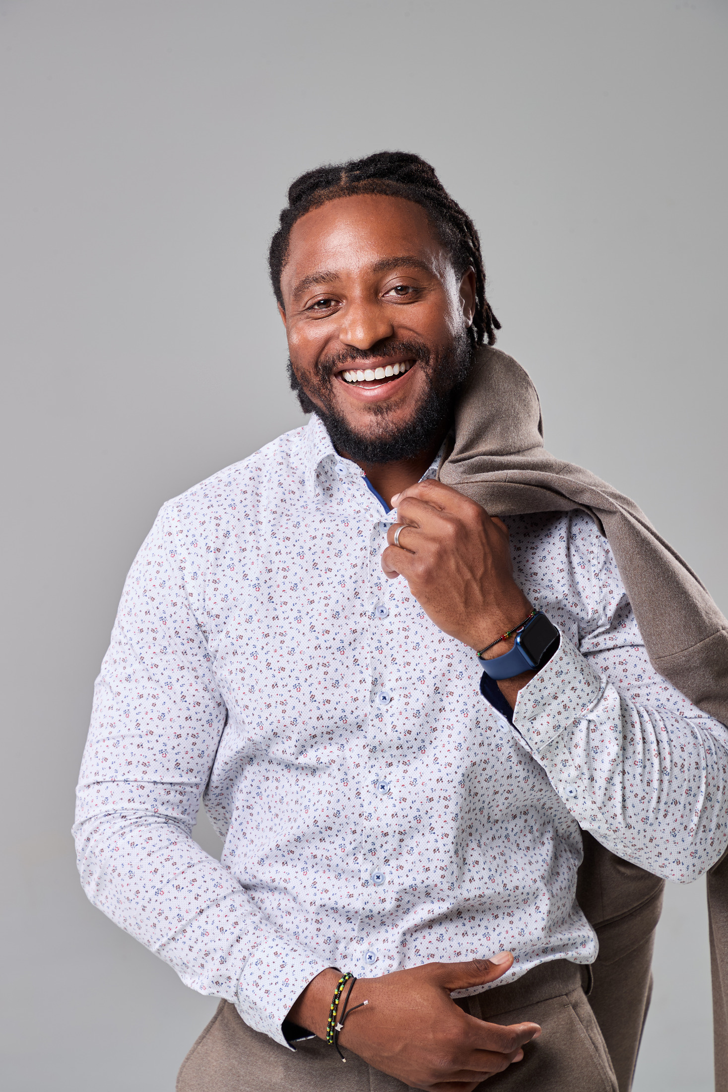 Man in a patterned shirt smiling with a jacket over his shoulder, grey studio portrait