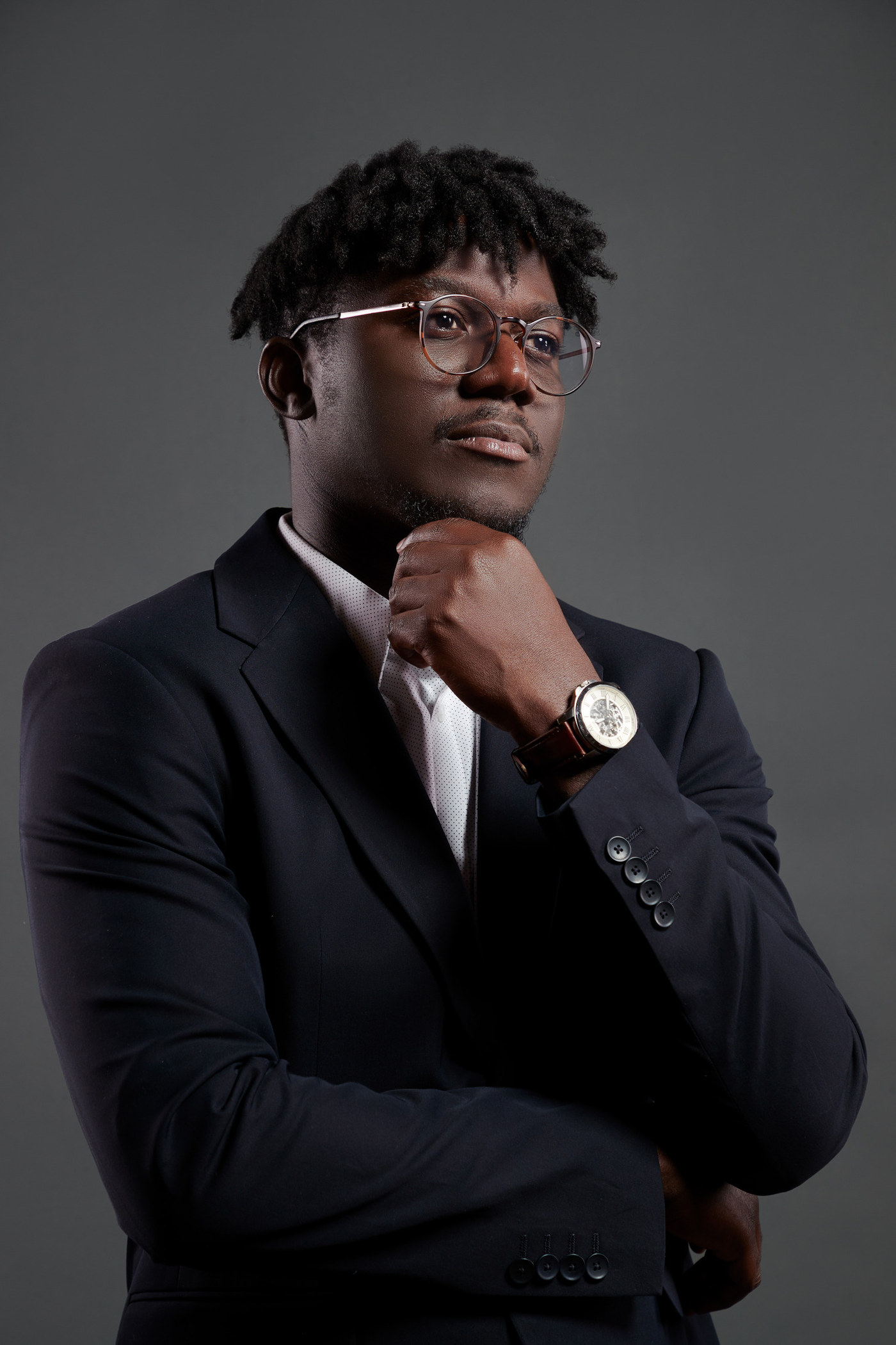 Executive portrait of a man in a navy blazer and round glasses, hand at chin, on a grey studio backdrop